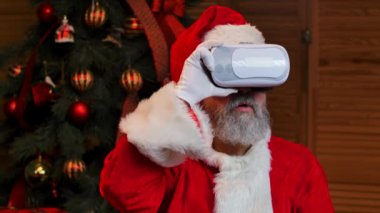 Noel Baba 'nın sanal gerçeklik gözlüğü takan portresi. Yüzünde hayret verici bir ifade var. Kırmızı takım elbiseli ve şapkalı yaşlı adam Noel ağacının arka planında. Kapatın. Yavaş çekim.