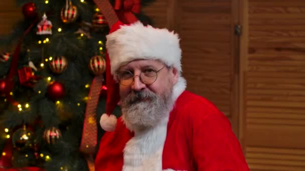 Porträtt av jultomten som dansar konstigt. Gamling med skägg i röd kostym och hatt mot bakgrund av julgran med leksaker och ljus. Glad nyårskväll. Närbild. Långsamma rörelser. — Stockvideo