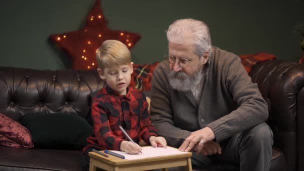 El niño dibuja un dibujo festivo con bolígrafos de fieltro. El abuelo y el nieto se sientan en un sofá en una habitación decorada cerca de un árbol de Navidad brillante. Concepto de vacaciones familiares. Nochebuena. Movimiento lento. — Vídeos de Stock