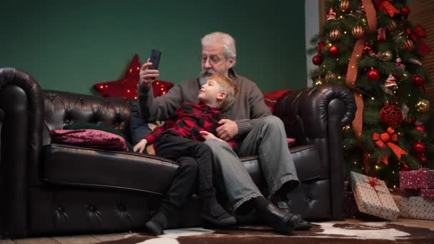 エレガントな老人と小さな男の子がスマートフォンでビデオ通信について話しています。祖父と孫は輝くクリスマスツリーの近くの装飾された部屋のソファに座っています。スローモーション. — ストック動画