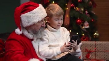 Noel Baba ve küçük bir çocuk akıllı telefona tıklayarak internetten bir Noel hediyesi seçiyor. E ticaret. Yeni yıl ruhu, tatiller ve kutlamalar kavramı. Kapatın. Yavaş çekim.