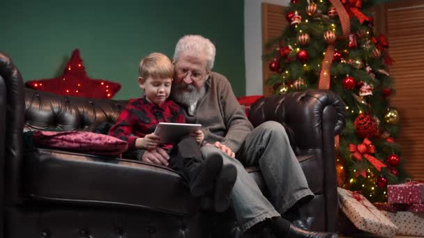 Eleganter alter Mann und kleiner Junge im Gespräch über Videokommunikation auf dem Tablet. Großvater und Enkel sitzen auf einem Sofa in einem geschmückten Raum neben einem leuchtenden Weihnachtsbaum. Zeitlupe. — Stockvideo