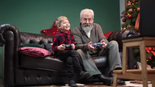 Viejo abuelo feliz con nieto jugando con joysticks. Elegante hombre de pelo gris con un niño pequeño está sentado en un sofá en una habitación decorada cerca de un árbol de Navidad brillante. Movimiento lento — Vídeos de Stock