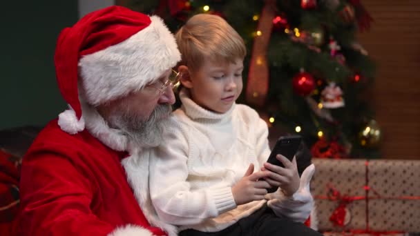 산타 클로스와 어린 남자 아이가 스마트폰을 클릭하고 인터넷에서 크리스마스 선물을 고르고 있습니다. 전자 상거래. 새해 정신의 개념, 휴일 과 축하 행사. 닫아. 느린 동작. — 비디오