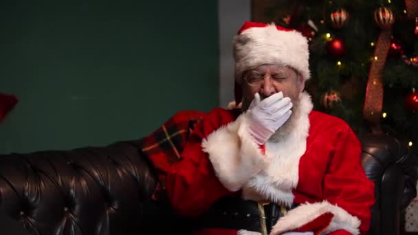 Le Père Noël fatigué bâille et s'endort assis sur le canapé. Vieil homme avec barbe en costume rouge et un chapeau dans une pièce décorée près d'un sapin de Noël lumineux et des boîtes avec des cadeaux. Ferme là. Mouvement lent. — Video