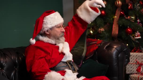 Le Père Noël fatigué bâille et s'endort assis sur le canapé. Vieil homme avec barbe en costume rouge et un chapeau dans une pièce décorée près d'un sapin de Noël lumineux et des boîtes avec des cadeaux. Ferme là. Mouvement lent. — Video