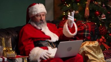 Şirin Noel Baba bilgisayar kullanarak iletişim kuruyor. Kırmızı takım elbiseli ve şapkalı yaşlı adam parlak Noel ağacının yanındaki süslü bir odada koltukta oturuyor. Kapatın. Yavaş çekim.