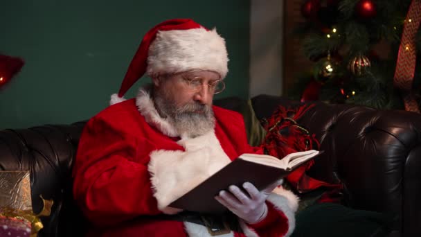 De Kerstman leest een boek met sprookjes. Oude man met een baard in rood pak en hoed zit op de bank in versierde kamer in de buurt van gloeiende kerstboom en dozen met geschenken. Sluit maar af. Langzame beweging. — Stockvideo