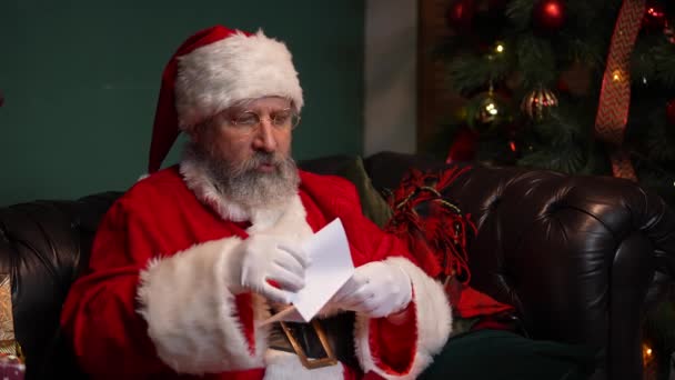 Santa Claus čte dopisy s radostí a s úsměvem na tváři. Starý muž v červeném obleku a klobouku sedí na pohovce ve zdobeném pokoji poblíž zářícího vánočního stromku a dárků. Zavřít. Zpomalený pohyb. — Stock video