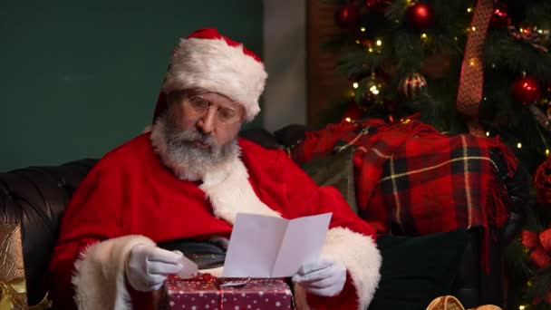 산타클로스는 편지를 읽고 아이들에게 줄 선물을 선택 한다. 빨간 양복을 입고 모자를 쓴 노인이 빛나는 크리스마스 트리와 선물 상자 옆에 장식 된 방에 있는 소파에 앉아 있다. 닫아. 느린 동작. — 비디오