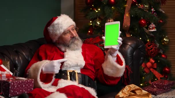De Kerstman ligt op de bank in de buurt van een versierde kerstboom en toont een tablet met een groen scherm chromen sleutel, terwijl het maken van een duim omhoog gebaar. Nieuwjaarsreclame, promotie. Sluit maar af. Langzame beweging. — Stockvideo