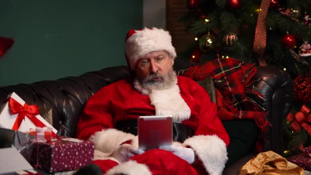 Санта-Клаус общается по видеосвязи с помощью планшета. Старик в красном костюме и шляпе лежит на диване в украшенной комнате возле светящейся елки и коробок с подарками. Закрывай. Медленное движение. — стоковое видео