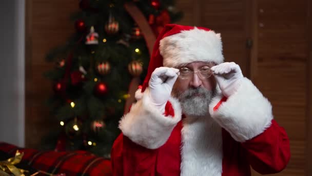 Retrato de Santa Claus mira a la cámara en serio, guiña y sonríe. Anciano con barba en traje rojo y sombrero sobre el fondo del árbol de Navidad con juguetes y luces. Movimiento lento. — Vídeo de stock