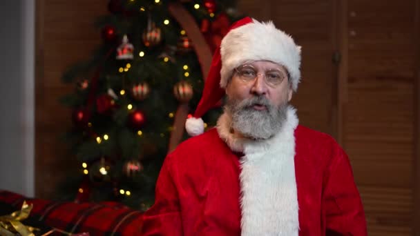 Portret van de Kerstman die naar de camera kijkt en iedereen feliciteert met de feestdagen. Oude man met baard in rood pak en hoed tegen de achtergrond van de kerstboom. Gelukkig Nieuwjaar. Langzame beweging — Stockvideo