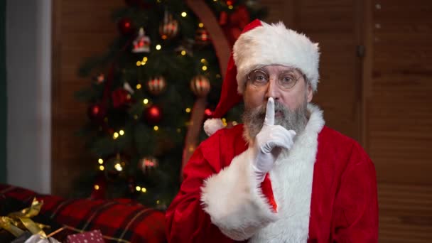 Портрет Санта-Клауса, задумчиво оглядывающийся вокруг, делает тайный жест и подмигивает. Старик с бородой в красном костюме и шляпе на фоне рождественской елки с игрушками и огнями. Медленное движение. — стоковое видео