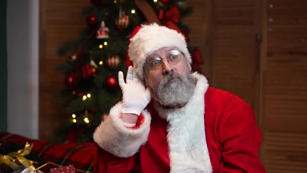 Портрет Деда Мороза, держащего руку за ухо, пытающегося слушать интересные новости. Старик в красном костюме и шляпе на фоне елки с игрушками и огнями. Медленное движение. — стоковое видео