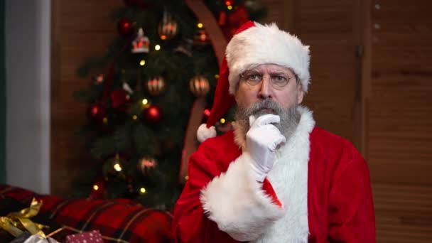 Portrait of Santa Claus looking pensively around, makes secret gesture and winks. Old man with beard in red suit and hat against the background of Christmas tree with toys and lights. Slow motion. — Stock Video