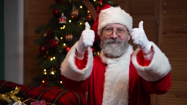 Portret van de Kerstman die in de camera kijkt en duimen laat zien als gebaar. Oude man met baard in rood pak en hoed tegen de achtergrond van kerstboom met speelgoed en verlichting. . Langzame beweging — Stockvideo