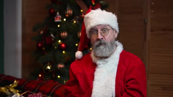 Portrait du Père Noël regardant la caméra écoute attentivement et fait un geste ok. Vieil homme en costume rouge et chapeau sur le fond de l'arbre de Noël avec des jouets et des lumières. Mouvement lent. — Video