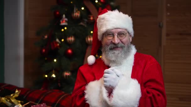 Retrato de Santa Claus mirando a la cámara escucha atentamente y hace un gesto pulgar hacia arriba. Hombre viejo en traje rojo y sombrero sobre el fondo del árbol de Navidad con juguetes. Movimiento lento. — Vídeos de Stock