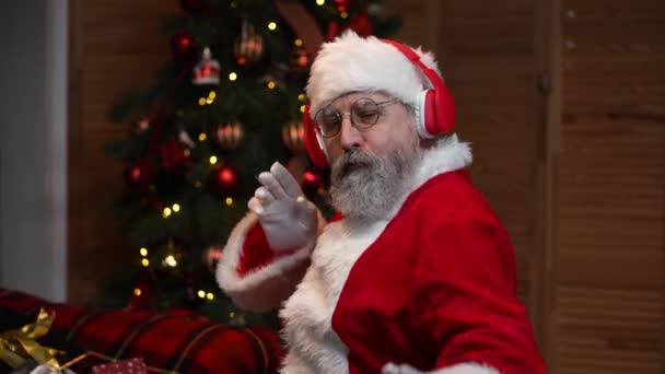 Портрет Санта Клауса, танцующего смешно в больших красных наушниках. Старик с бородой в красном костюме и шляпе на фоне рождественской елки с игрушками и огнями. Счастливого Нового Года. Медленное движение. — стоковое видео