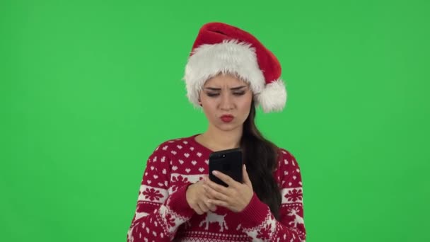 Портрет милой девушки в шляпе Санта-Клауса сердито отправляет смс на телефон. Зеленый экран — стоковое видео