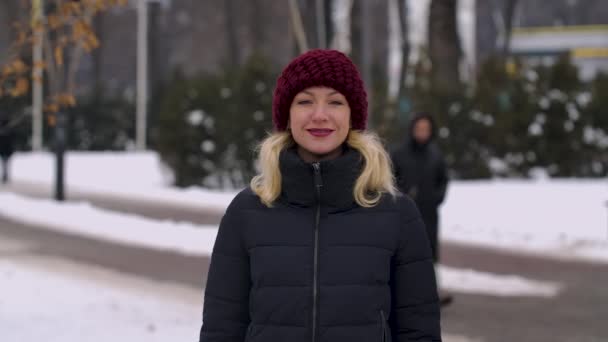 Retrato de uma menina sorridente em pé no meio de um parque da cidade em roupas de inverno. A mulher gosta do tempo de inverno, respirando no ar gelado. O fundo está desfocado. Movimento lento. Fechar. — Vídeo de Stock