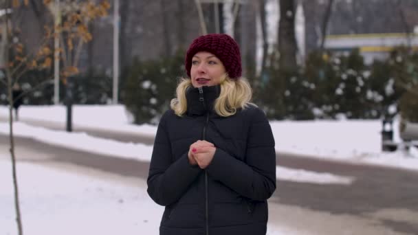 Kobieta w zimowych ciuchach stoi w środku parku w mroźny dzień. Dziewczyna zapomniała rękawiczek i rozgrzewa ręce, oddychając nimi i pocierając nimi o siebie. Zwolnij trochę. Zamknij się.. — Wideo stockowe