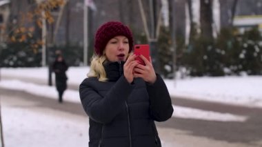 Mutlu bir kızın portresi bir kış şehrinin caddesinde akıllı bir telefonla selfie çekiyor. Modern teknoloji, yaşam tarzı ve neşeli insanlar konsepti. Kapatın. Yavaş çekim.
