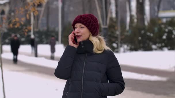 En ung charmig kvinna med blont hår talar på en mobiltelefon, stående mitt på gatan. Vintertid i staden. Bakgrunden är suddig. Sakta i backarna. Närbild. — Stockvideo