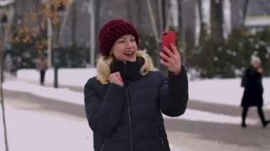 Kışın karlı bir parkta elinde akıllı bir telefonla konuşan bir kadın. Kız, müdahaleciyi selamlar, baş parmağını kaldırır, hava öpücüğü gönderir ve elini sallar. Kapatın. Yavaş çekim.