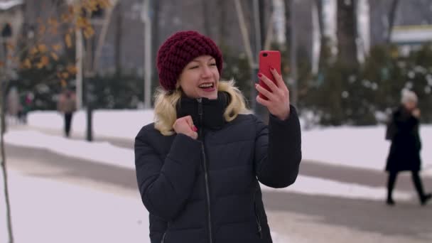 Жінка в зимовому сніжному парку тримає смартфон і розмовляє на відеодзвінок. Дівчина вітає співрозмовника, показує жест на великий палець, посилає повітряний поцілунок і хвилює її руку. Близько. Повільний рух . — стокове відео