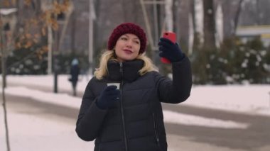 Akıllı telefonuyla selfie çeken bir kızın portresi bir fincan kahveyle, kış şehrinin sokaklarında çay içerken. Kız alınan fotoğraflara bakıyor. Kapatın. Yavaş çekim.