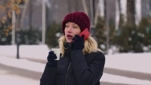 Mujer joven hablando por celular y bebiendo café aromático caliente de una taza de papel. Una chica se encuentra en medio de un parque de invierno cubierto de nieve. El fondo está borroso. En cámara lenta. De cerca.. — Vídeo de stock