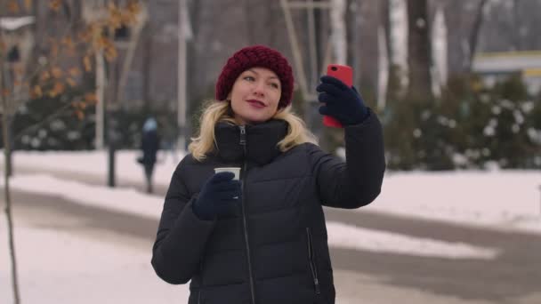 Akıllı telefonuyla selfie çeken bir kızın portresi bir fincan kahveyle, kış şehrinin sokaklarında çay içerken. Kız alınan fotoğraflara bakıyor. Kapatın. Yavaş çekim. — Stok video