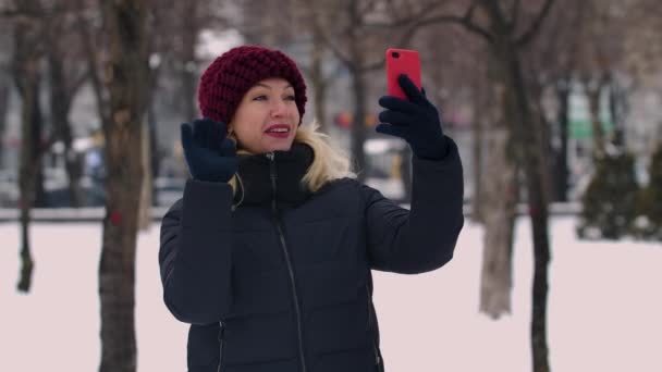 Mujer en un parque nevado de invierno sosteniendo un teléfono inteligente y hablando en una videollamada. La chica saluda al interlocutor, muestra un gesto de pulgar hacia arriba, envía un beso aéreo y agita la mano. De cerca. Movimiento lento. — Vídeo de stock