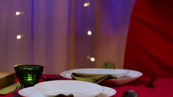 Žena v červených šatech umístí dvě sklenice šampaňského na slavnostní stůl vedle talířů a příborů. Home útulný pokoj, zdobené na oslavu Nového roku nebo Vánoc. Ruce k sobě. Zpomalený pohyb. — Stock video