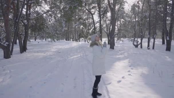 Dívka v zimě po kabátě kráčí po stezce v zasněženém zimním lese. Mladá žena se rozhlíží s potěšením a úsměvy. V lese je hodně sněhu. Sněžná zima. Slunečný mrazivý den. Zpomalený pohyb. — Stock video