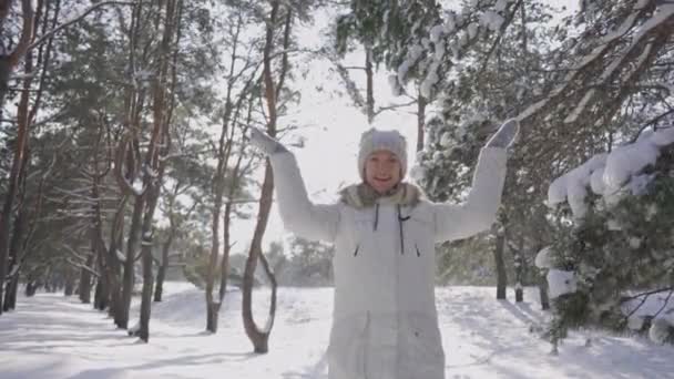 Jong meisje verzamelt sneeuw in haar handen en geeft het over. Witte pluizige sneeuw schittert in zonnestralen. Vrouw wordt gespeeld met sneeuw in besneeuwd dennenbos op zonnige dag. Prachtige winternatuur. Langzame beweging. — Stockvideo