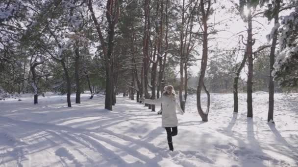 Fille en vêtements d'hiver et mitaines chaudes marche à travers une forêt de pins enneigés et est très heureux. Une femme souriante jette de la neige scintillante au soleil. Agréable promenade par une journée d'hiver glacée. Mouvement lent. — Video