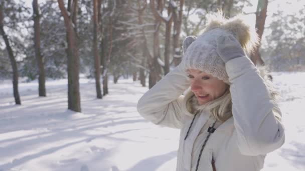 Πορτρέτο του χαριτωμένο κορίτσι σε χειμωνιάτικα ρούχα συνθλίβει το χιόνι από το καπέλο και ζεσταίνει τα χέρια σε ζεστά γάντια. Το κορίτσι απολαμβάνει παγωμένη ηλιόλουστη μέρα. Όμορφη χειμωνιάτικη φύση καλυμμένη με χιόνι. Κλείσε. Αργή κίνηση. — Αρχείο Βίντεο