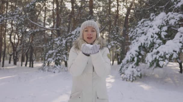 Happy cute girl, dressed in warm winter clothes, blows off snow from her hands to camera, outdoors, in snowy park or forest. Beautiful blurred background of snowy nature. Slow motion. Close up. — Stock Video