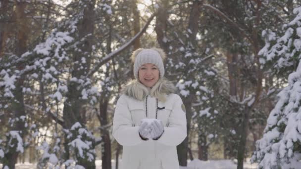 Szczęśliwy cute girl w zimie ubrania rzuca śnieg i śmieje się wesoło. Śnieżna zimowa zabawa w słoneczny dzień. Piękne rozmazane tło śnieżnej przyrody w sosnowym lesie. Zwolnij trochę. Zamknij się.. — Wideo stockowe