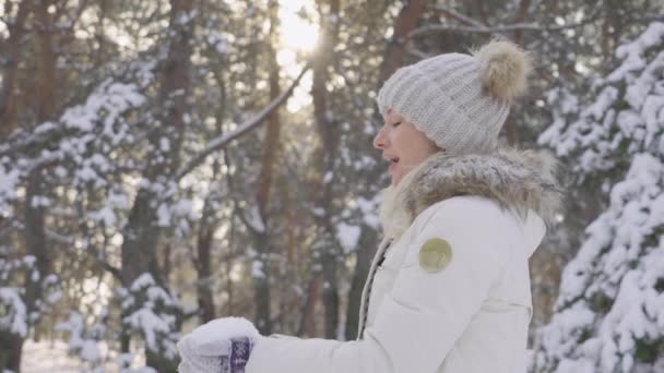 Вид сбоку симпатичная девушка в теплой зимней одежде бросает снег и весело смеется. Красивый размытый фон снежной природы в сосновом лесу. Медленное движение. Закрыть. — стоковое видео