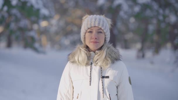 Porträtt av en söt flicka i varma vinterkläder tittar på kameran och ler. Ung kvinna poserar utomhus på en suddig bakgrund av en snöig skog. Närbild. Långsamma rörelser. — Stockvideo