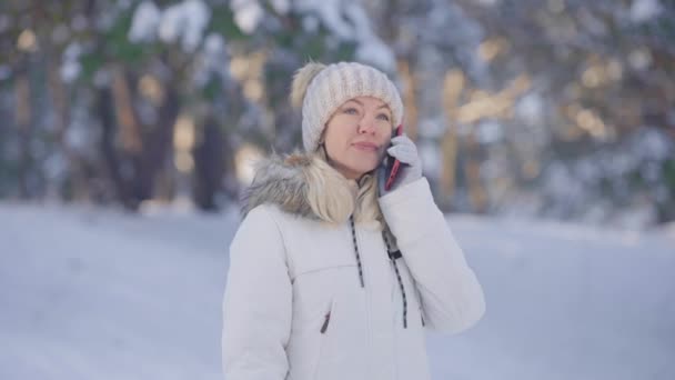 Portrait of cute girl in warm winter clothes talking for mobile phone and rejoice. Young woman posing outdoors on blurred background of snowy forest. Close up. Slow motion. — Stock Video