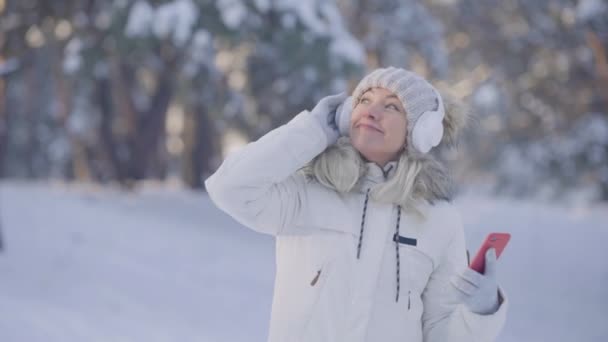 Retrato de una linda chica en ropa de invierno caliente escuchando música en grandes auriculares blancos usando un teléfono inteligente. Mujer joven posando al aire libre sobre un fondo borroso de bosque nevado. De cerca. Movimiento lento. — Vídeo de stock