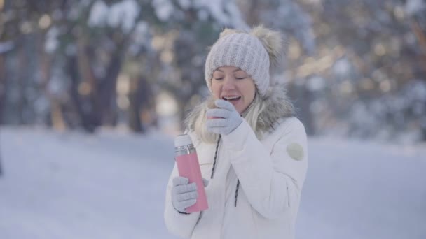 Портрет милой девушки в теплой зимней одежде пьет горячий чай из термоса. Молодая женщина позирует на открытом воздухе на размытом фоне снежного леса. Закрывай. Медленное движение. — стоковое видео