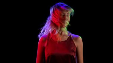 Genç, güzel sarışın, saçlarını sallıyor ve kameraya cilveli bir şekilde bakıyor. Siyah stüdyo arka planında neon ışıklarla poz veren kız model. Kapatın. Yavaş çekim.