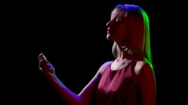 Üzerine sprey şişeden parfüm serpiştiren genç ve güzel bir sarışın siluetinin yan görüntüsü. Siyah stüdyo arka planında neon ışıklarla poz veren sevimli bir kızın portresi. Kapatın. Yavaş çekim.